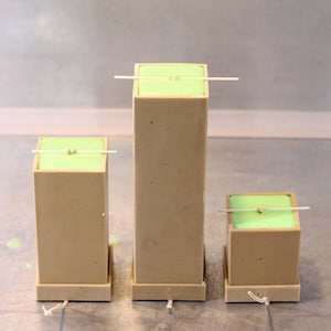 May include: Three rectangular candles with wicks, each with a light green top and a beige base. The candles are different sizes and are sitting on a table.