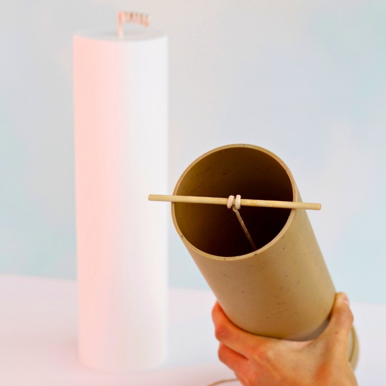 May include: A brown cylindrical container with a white wick and a wooden stick across the top. The container is being held by a hand.
