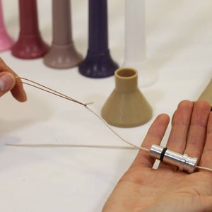 May include: A close-up of a hand holding a silver metal spool with a white string attached to it. The string is also attached to a brown plastic cone. The cone is sitting on a white surface.