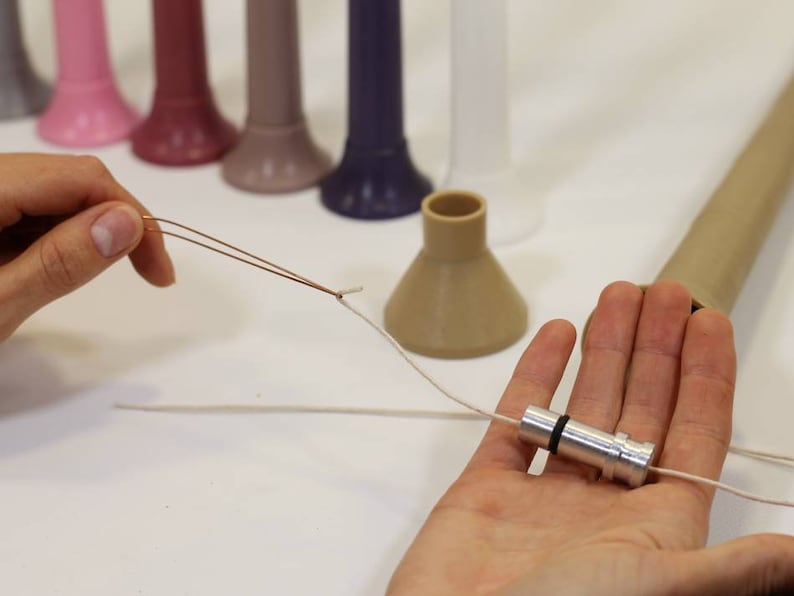 May include: A close-up of a hand holding a silver metal spool with a white string attached to it. The string is also attached to a brown plastic cone. The cone is sitting on a white surface.