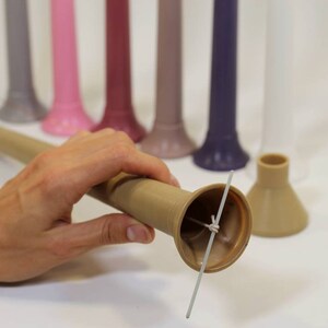 May include: A close-up of a brown candle with a white wick. The candle is being held by a hand and is being inserted into a brown candle holder. The candle holder has a wide base and a narrow top. There are several other candles in the background, each a different color.
