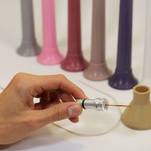 May include: A silver metal candle wick holder with a copper wire attached. The holder is being held in a hand. There are several colorful candle holders in the background.