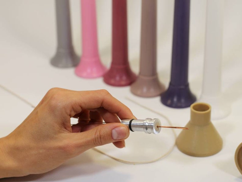 May include: A silver metal candle wick holder with a copper wire attached. The holder is being held in a hand. There are several colorful candle holders in the background.
