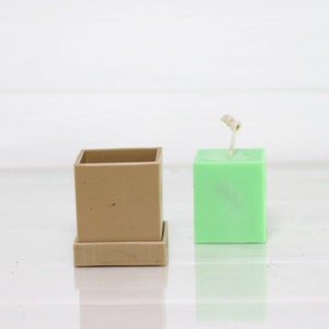 May include: A square, light green candle with a wick sticking out of the top. The candle is sitting next to a square, brown planter with a removable base.