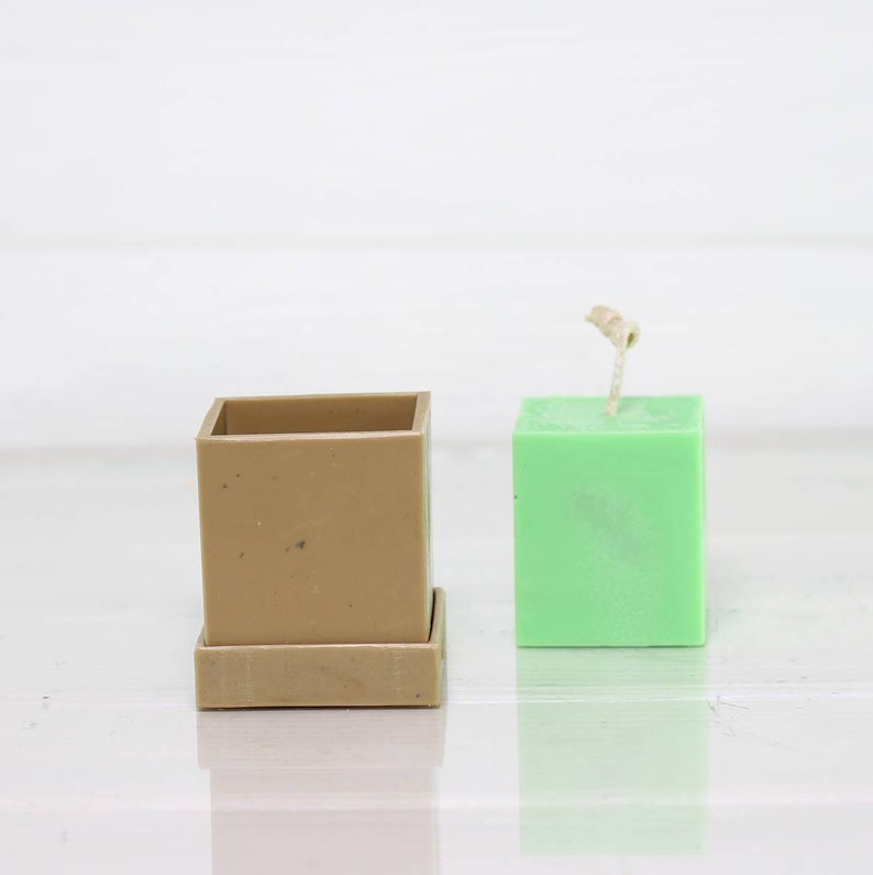 May include: A square, light green candle with a wick sticking out of the top. The candle is sitting next to a square, brown planter with a removable base.