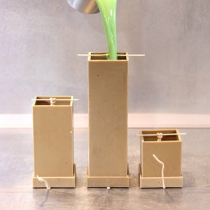 May include: Three rectangular candle molds, two smaller and one larger, with wicks inserted. Green wax is being poured into the largest mold.