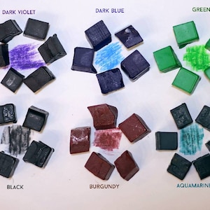 May include: Nine watercolor paint pans in various colors, including dark violet, dark blue, green, black, burgundy, and aquamarine. Each pan is arranged in a circle with the color name printed above it.