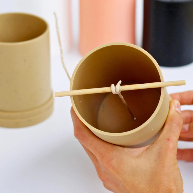 May include: A close-up of a beige candle jar with a wooden stick and a white string running through it. The jar is being held by a hand.