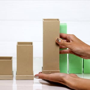 May include: Three beige square-shaped containers with a base, arranged in ascending order of size. The largest container is being held by a hand, and there are three green foam blocks behind it.