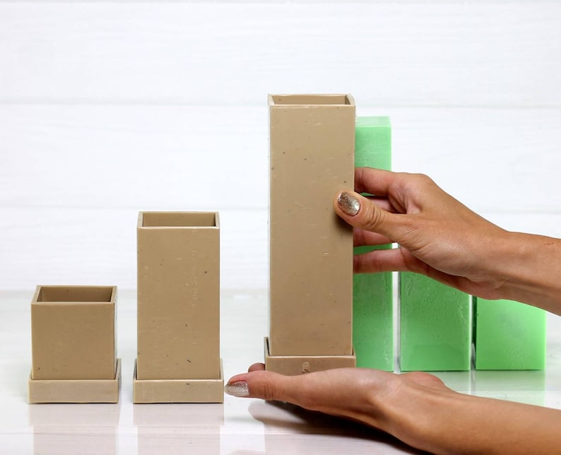 May include: Three beige square-shaped containers with a base, arranged in ascending order of size. The largest container is being held by a hand, and there are three green foam blocks behind it.
