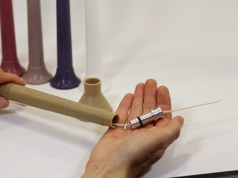 May include: A close-up of a hand holding a silver candle wick holder with a white wick threaded through it. The holder is made of metal and has a black rubber ring around the top. The wick is about 10 inches long.