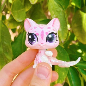 Può includere: Una statuina a forma di gatto rosa con un motivo a macchie e grandi occhi espressivi. Il gatto giocattolo ha una lunga coda curva ed è ambientato su uno sfondo di foglie verdi. La statuina è un giocattolo da collezione.