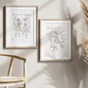 Puede incluir: Dos impresiones art&iacute;sticas enmarcadas con ilustraciones de figuras con vestidos fluidos y detalles florales. Las impresiones tienen texto rojo y est&aacute;n sobre un fondo neutro. La obra de arte est&aacute; en un marco de madera clara.