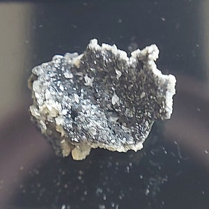 May include: A small, irregularly shaped piece of dark grey rock with white crystals embedded in it.