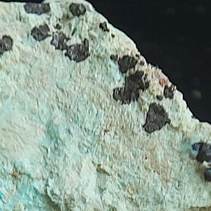 May include: A close-up of a light blue and white mineral specimen with black and brown inclusions. The mineral has a rough, textured surface.
