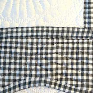 Op de afbeelding: Close-up van een zwart-witte gingham stof met een gestikt ontwerp. De stof is in een hoek van een groter stuk stof genaaid.