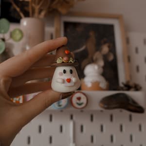 Op de afbeelding: Een klein wit spookfiguurtje met een bruine hoed met een rood hart en een rood hart in zijn hand.