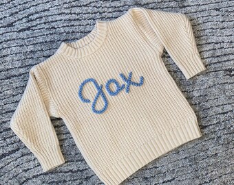 May include: A cream coloured knitted sweater with a blue embroidered name "Jax" on the front.