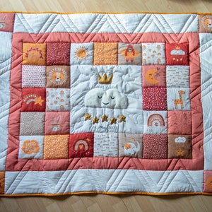 Könnte beinhalten: Eine handgefertigte Babydecke mit Patchwork-Design. Die Decke zeigt Quadrate mit verschiedenen gestickten Bildern, darunter eine Wolke, Sterne, ein Löwe und eine Giraffe. Die Farbpalette umfasst Orange, Weiß und Pfirsich. Die Decke hat einen weißen Rand und einen pfirsichfarbenen Rahmen.