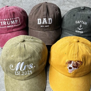 May include: Five different colored baseball caps with embroidered text. The caps are brown, red, green, yellow, and black. The text on the caps reads "DAD EST. 2022", "TRUMP 2024", "Mrs. EST. 2024", "CAPTAIN HERMAN", and a dog graphic.