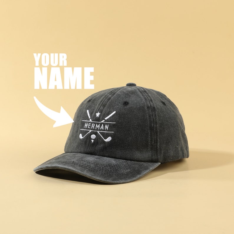 K&ouml;nnte beinhalten: Eine schwarze Baseballkappe aus Denim mit einem wei&szlig;en gestickten Design mit gekreuzten Golfschl&auml;gern und dem Wort "HERMAN". Der Text "YOUR NAME" steht &uuml;ber der Kappe.