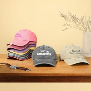 Puede incluir: Tres gorras de béisbol con el texto "Custom Embroidery" en ellas. Las gorras son de diferentes colores: rosa, gris y beige.