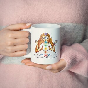 May include: White ceramic mug featuring a colorful illustration of a meditating figure with chakra symbols. The figure has long orange hair and is seated in a lotus position. The mug is held by a person wearing a pink sweater.