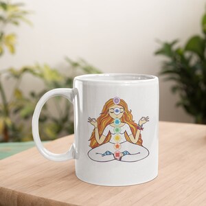 May include: White ceramic mug featuring a colorful illustration of a meditating figure with chakra symbols. The figure has long orange hair and is seated in a lotus position. The mug has a curved handle and is set on a wooden surface.