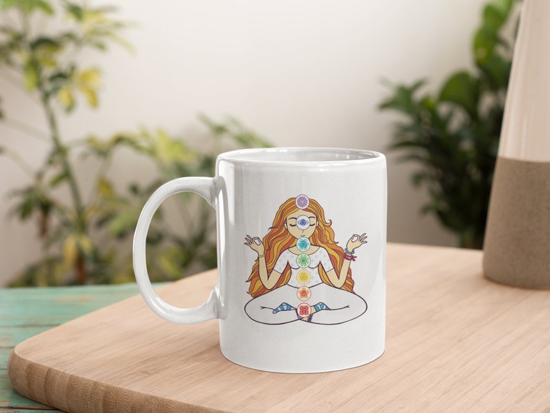 May include: White ceramic mug featuring a colorful illustration of a meditating figure with chakra symbols. The figure has long orange hair and is seated in a lotus position. The mug has a curved handle and is set on a wooden surface.
