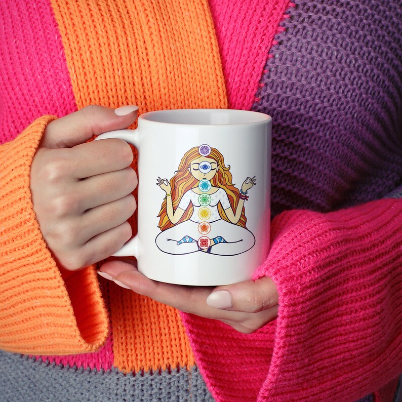 May include: White ceramic mug featuring a seated woman with long orange hair, eyes closed, and hands in a meditation pose. The design includes seven colorful chakra symbols aligned down the center of the woman's body. The mug is held by a person wearing a colorful sweater.