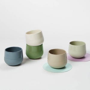 Könnte beinhalten: Vier Keramikbecher in verschiedenen Farben: Blau, Grün, Beige und Hellgrün. Die Becher sind übereinander gestapelt und stehen auf einer weißen Oberfläche.