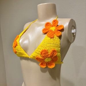 May include: A yellow crochet bikini top adorned with orange flower appliqués. The triangle top features a halter neck and is decorated with multiple orange flowers. The handmade top is displayed on a mannequin.