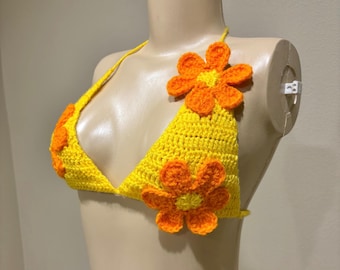 Puede incluir: Top de bikini de ganchillo amarillo adornado con aplicaciones de flores naranjas. El top triangular presenta un cuello halter y está decorado con múltiples flores naranjas. El top hecho a mano se exhibe en un maniquí.