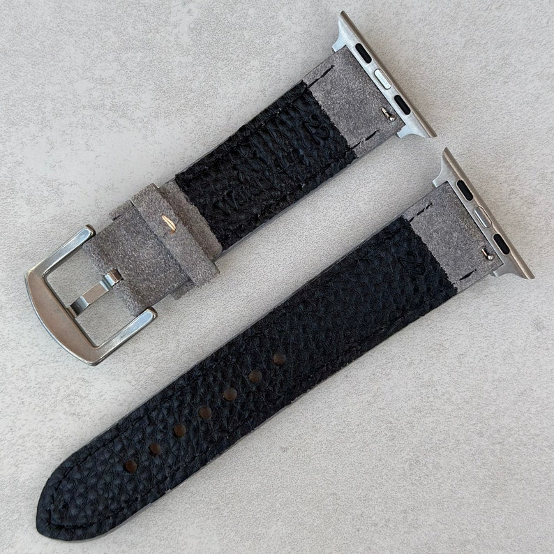 May include: Two Apple Watch bands, one gray and one black, made of leather with a textured finish. Both bands have silver metal adapters.