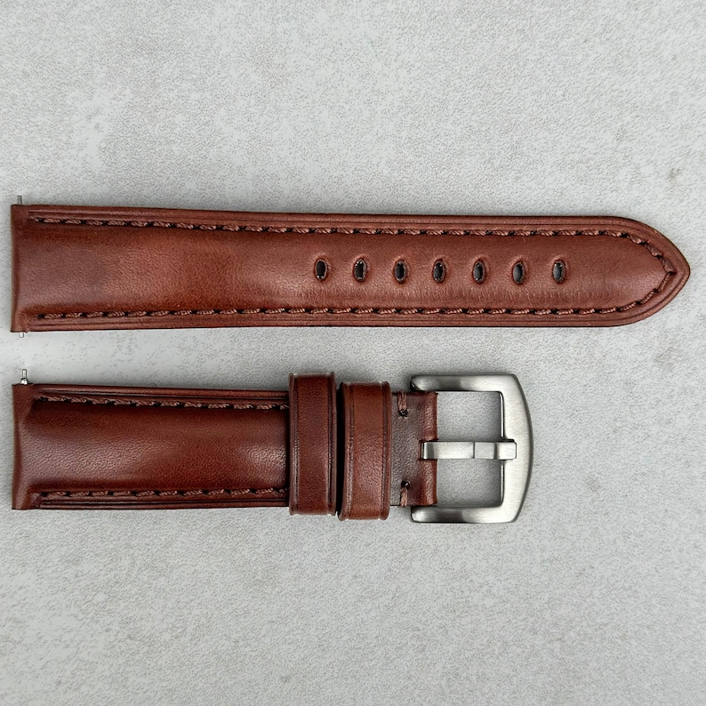 May include: A brown leather watch strap with a silver buckle. The strap has stitching around the edges and multiple holes for adjusting the size.