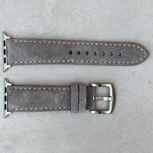 May include: Two gray suede watch bands with a silver buckle. The top band is a standard watch band, while the bottom band has a buckle that is designed for an Apple Watch.