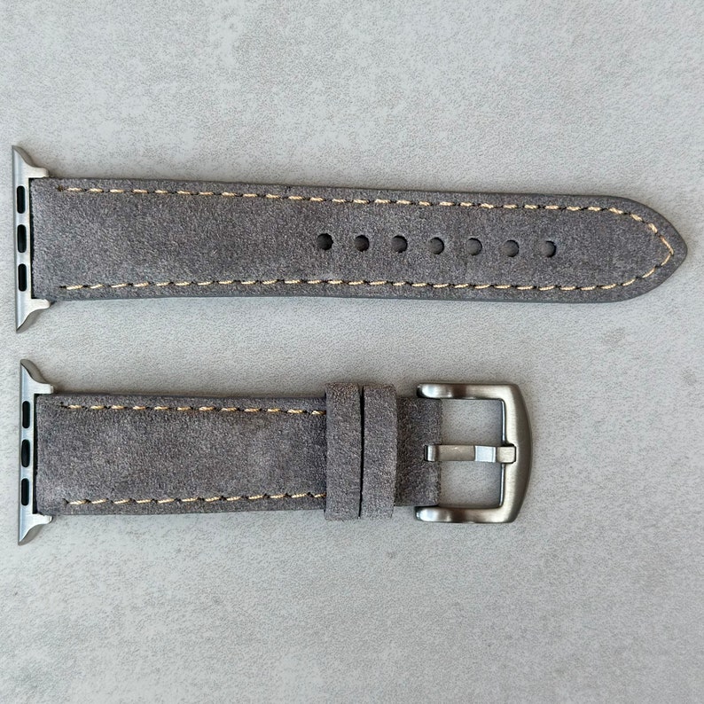 May include: Two gray suede watch bands with a silver buckle. The top band is a standard watch band, while the bottom band has a buckle that is designed for an Apple Watch.
