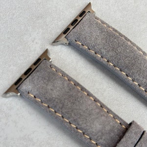 May include: Two gray suede watch bands with silver metal clasps and a contrasting yellow stitch. The bands are designed for Apple Watches.