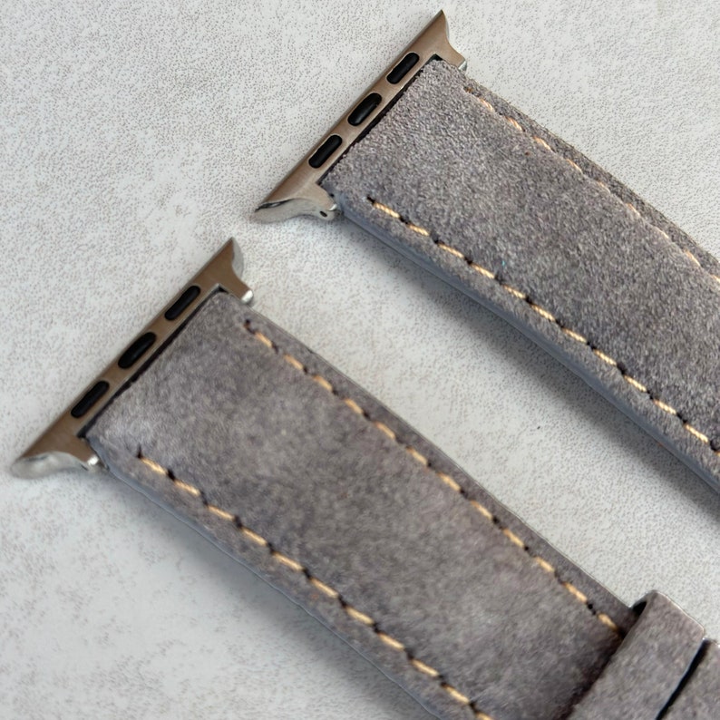 May include: Two gray suede watch bands with silver metal clasps and a contrasting yellow stitch. The bands are designed for Apple Watches.