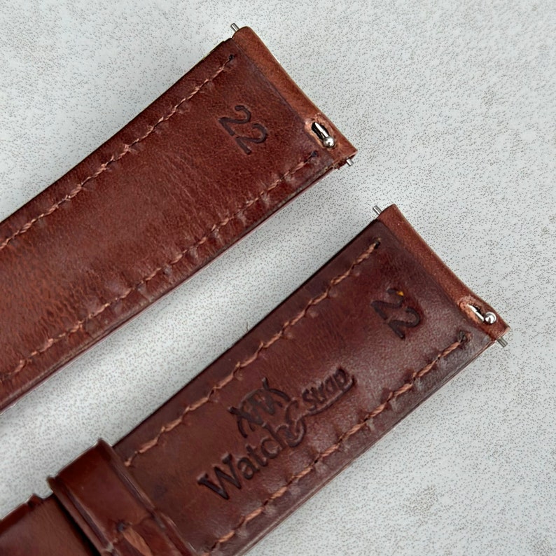 May include: Two brown leather watch straps with stitching. The strap on the top has the number "22" printed on it. The strap on the bottom has the text "Watch Strap" and the number "22" printed on it.