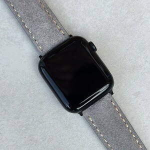 May include: A black Apple Watch with a gray leather band. The band has a light brown stitching detail.