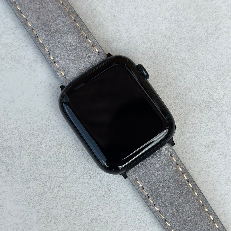 May include: A black Apple Watch with a gray leather band. The band has a light brown stitching detail.