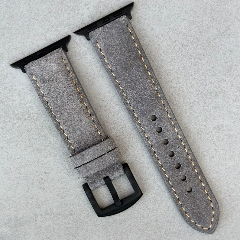 May include: A gray suede watch band with a black buckle and yellow stitching. The band is designed for an Apple Watch.