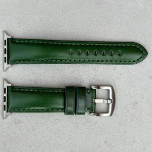 May include: A green leather watch strap with silver-coloured hardware. The strap is made of smooth, solid green leather and features a buckle closure. The strap is designed to fit a smartwatch.