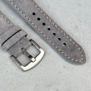 May include: A gray suede watch strap with a silver buckle. The strap has a light brown stitching detail.