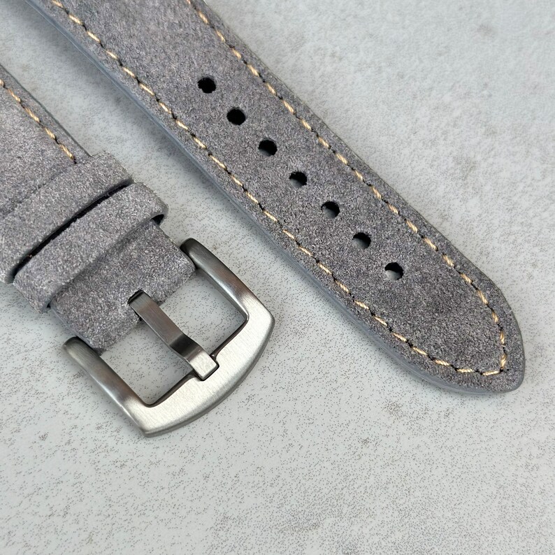 May include: A gray suede watch strap with a silver buckle. The strap has a light brown stitching detail.