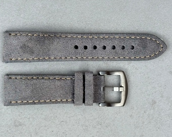 Può includere: Un cinturino per orologio in pelle scamosciata grigia con cuciture color crema. Il cinturino ha una fibbia color argento e un aspetto morbido e strutturato. Il cinturino è progettato per uno stile classico o vintage.