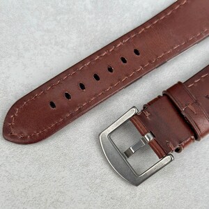 May include: A brown leather watch strap with a silver buckle. The strap has multiple holes for adjusting the fit.