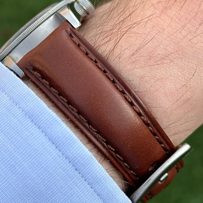May include: A brown leather watch strap with white stitching. The strap is attached to a silver watch buckle.