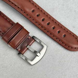 May include: A brown leather watch strap with a silver buckle. The strap has stitching along the edges and multiple holes for adjusting the size.